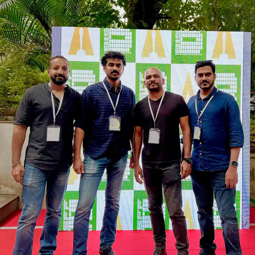 Four individuals pose together on a red carpet in front of a colorful backdrop featuring geometric patterns, surrounded by greenery.