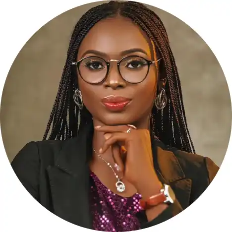 Close-up portrait of a confident woman with braided hair, wearing glasses and a stylish outfit, posing thoughtfully against a neutral background.