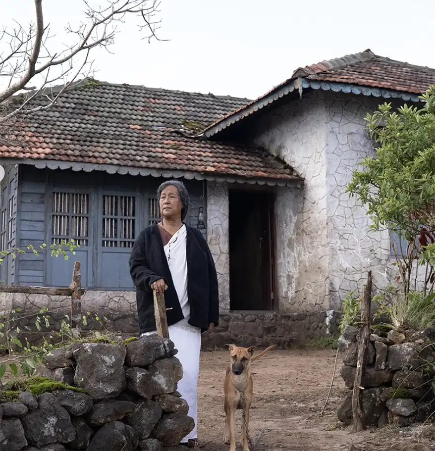 An older woman stands outside a rustic stone house, holding a stick and accompanied by a dog, amidst a natural setting.