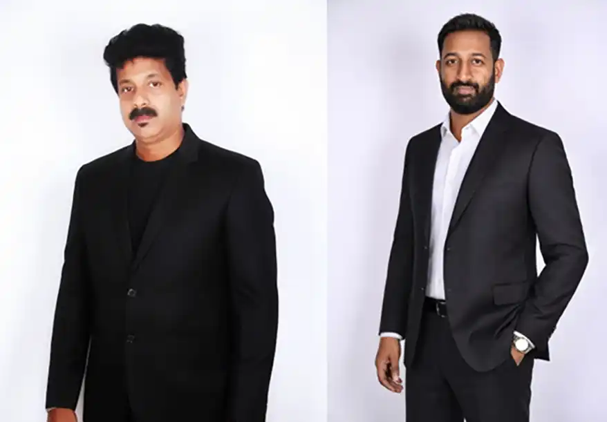 Two men in formal suits posing against a plain background. The man on the left wears a black suit with a black shirt, and the man on the right wears a black suit with a white shirt and has a beard.