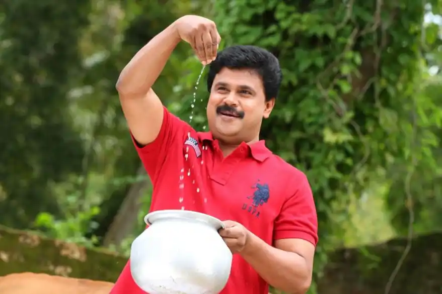 A man in a red shirt pouring liquid from a pot while smiling outdoors, surrounded by greenery.