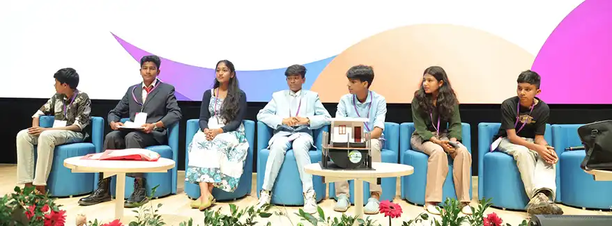 A group of young individuals seated on a stage during an event, with a variety of seating arrangements and floral decorations in the foreground.