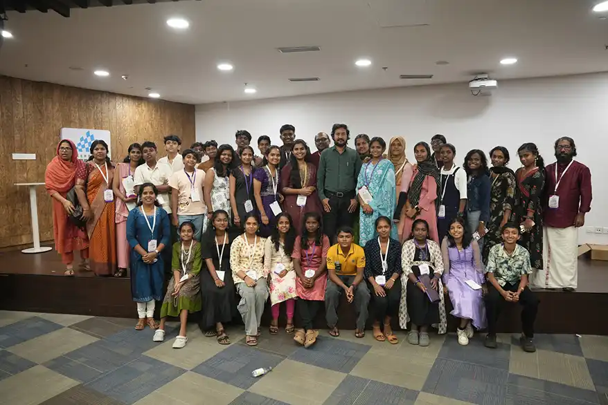 A large group photo of students and adults gathered in a conference room, smiling and posing together, with name tags visible on their attire.