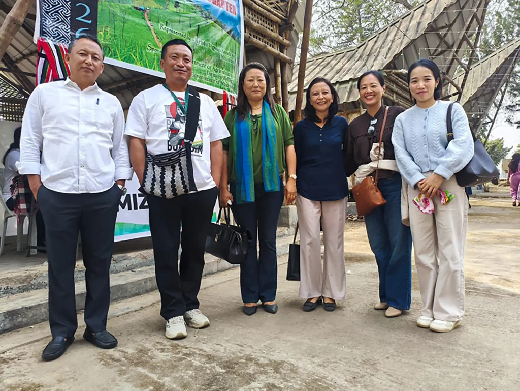 A group of six people standing outdoors in front of a wooden structure, with a banner in the background. They are dressed in casual attire and smiling at the camera.