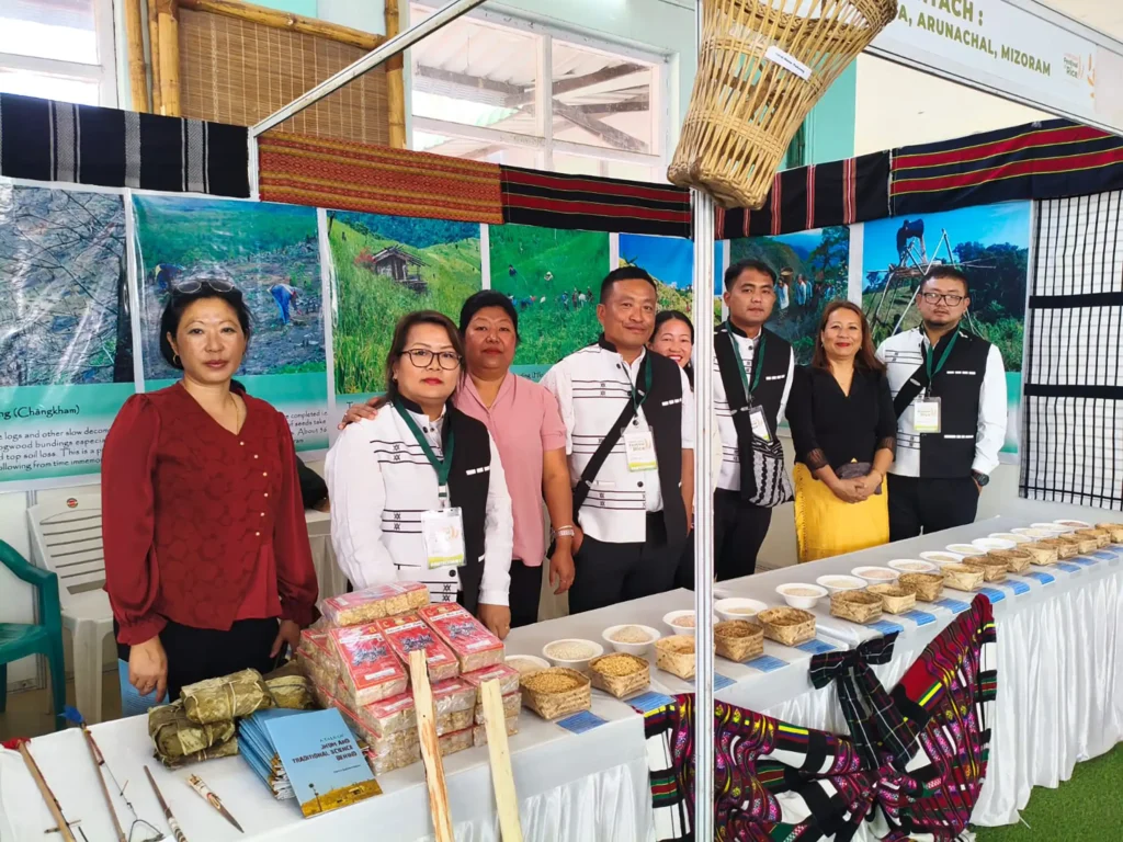 A group of seven individuals posing at a cultural exhibition booth, showcasing traditional crafts and food from Mizoram, Arunachal Pradesh, and other northeastern states of India. The booth features baskets, food samples in containers, and decorated with colorful fabric and photographs of local landscapes.