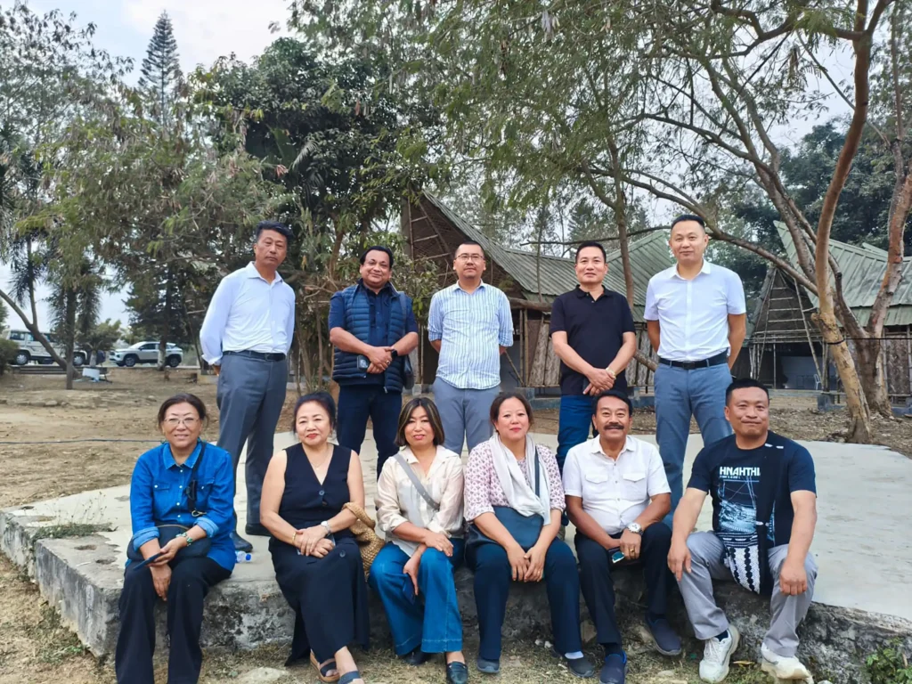 A group of twelve people posing for a photo outdoors, standing and sitting on a concrete surface with trees and buildings in the background.