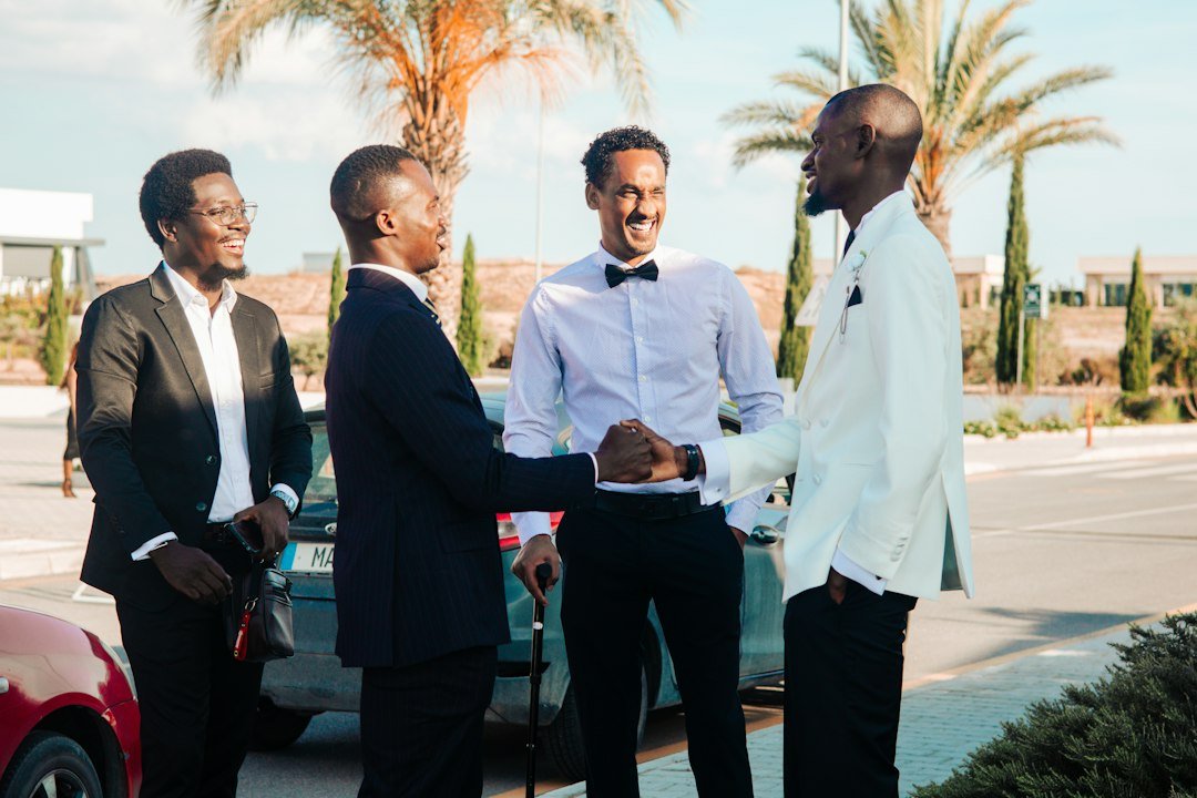 Four formally dressed men shaking hands outdoors with palm trees